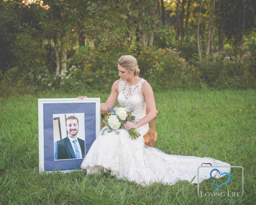 Woman in wedding dress sits on chair in grass beside picture frame of deceased husband