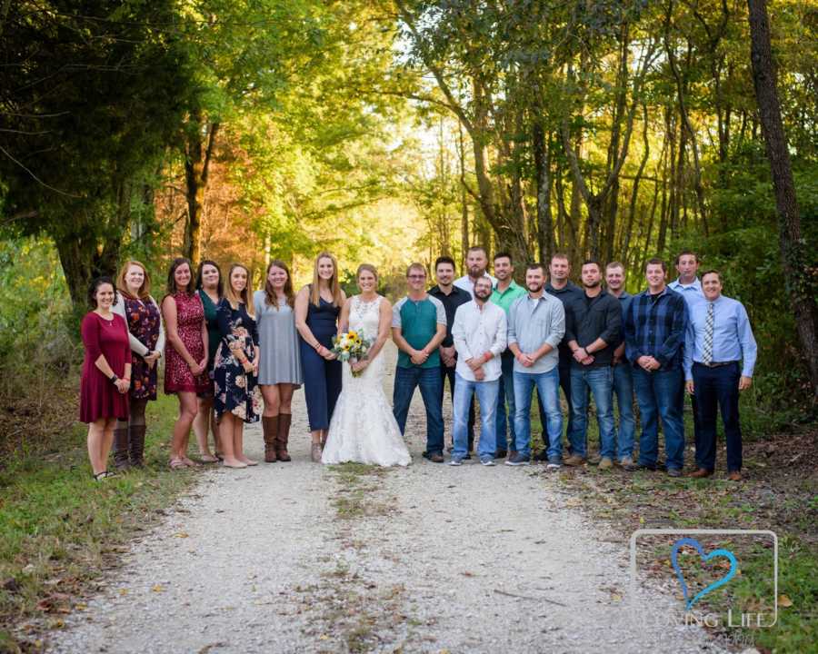 Woman in wedding gown stands on dirt path beside her family and late fiancee's family