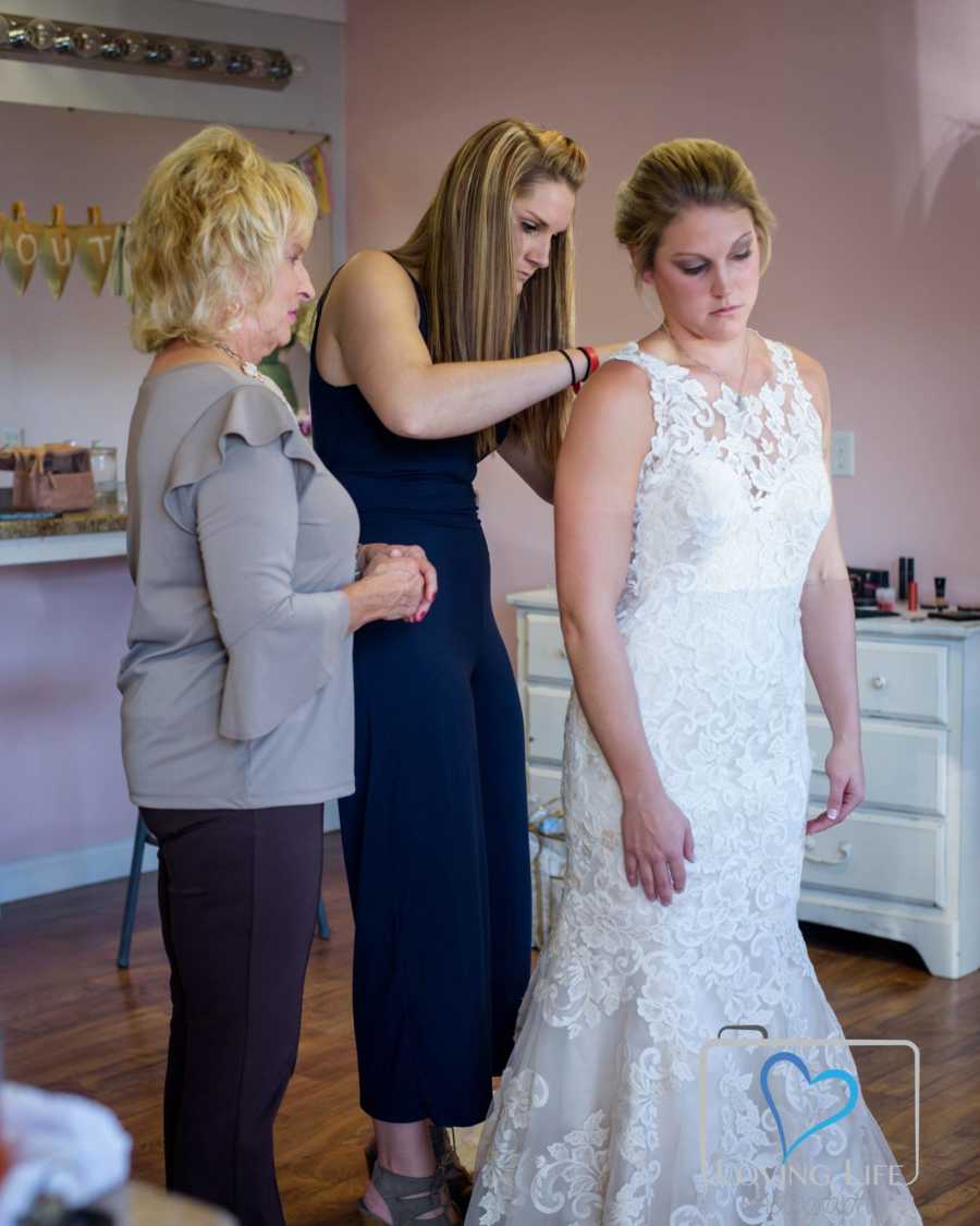 Woman stands in wedding dress as her sister buttons her up and mother watches