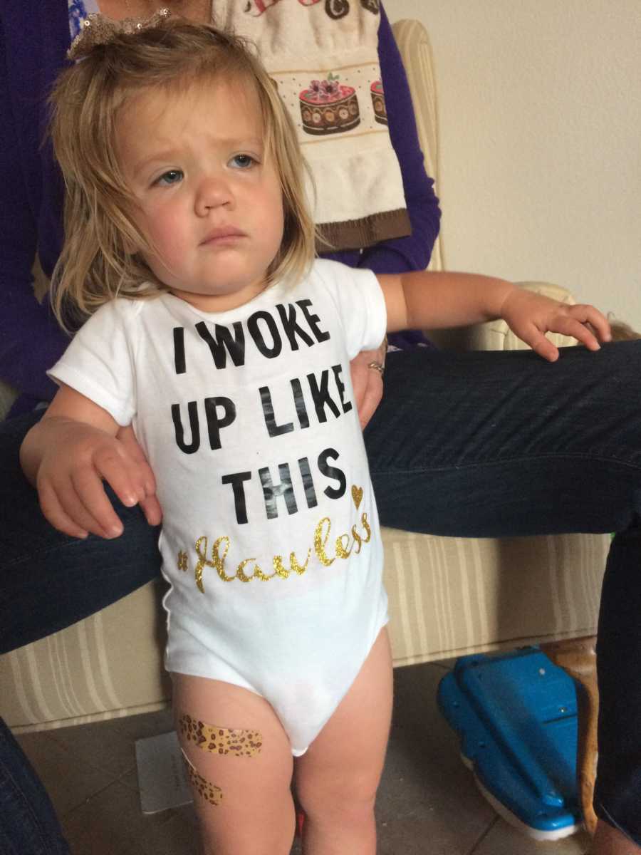 Toddler with sad face stands in white onesie that says, "I woke up like this #flawless"