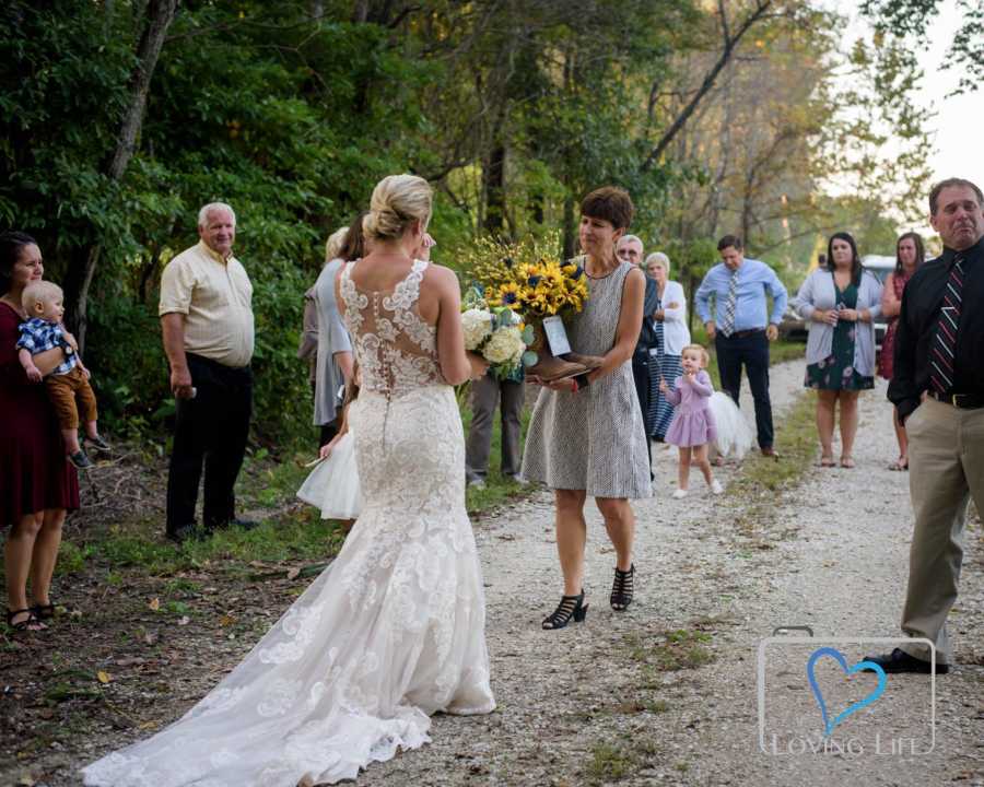 Bride walking down dirt path to meet late fiancee's mother who is holding his boots with flowers in them