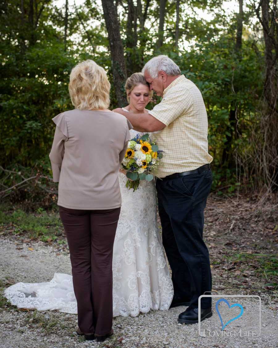 Woman is wedding gown is hugged by father as mother watches