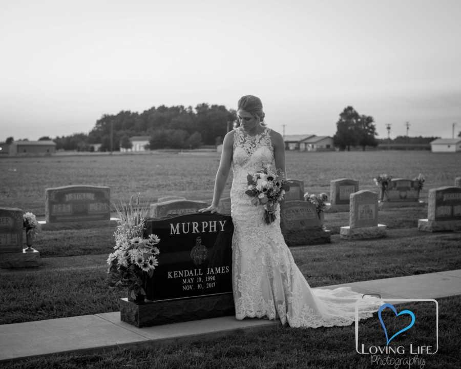 Woman in wedding gown stands beside late fiancee's head stone