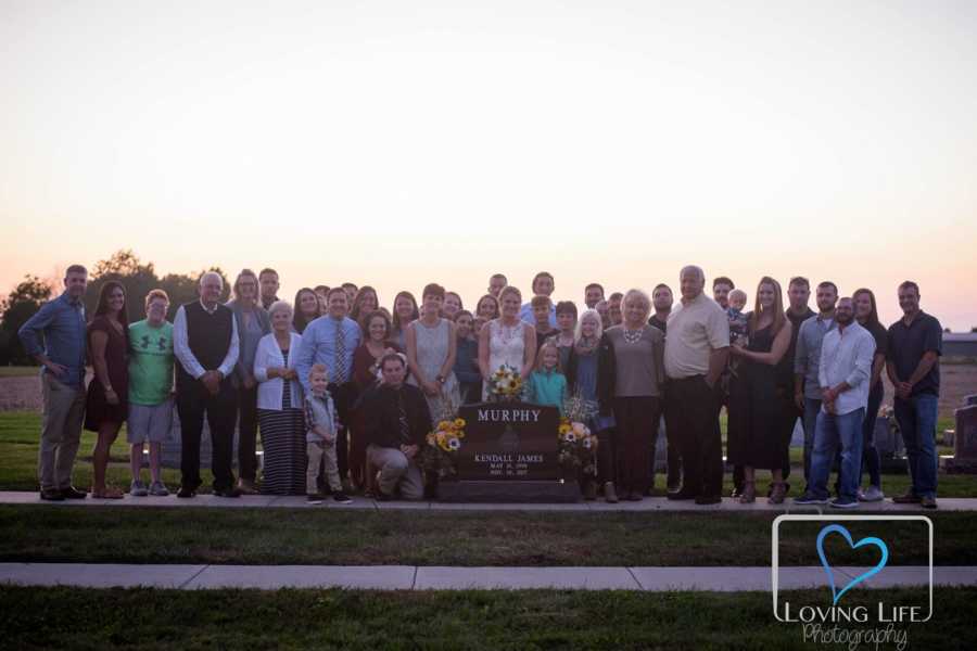 Woman in wedding gown stands behind late fiancee's headstone with his family and hers