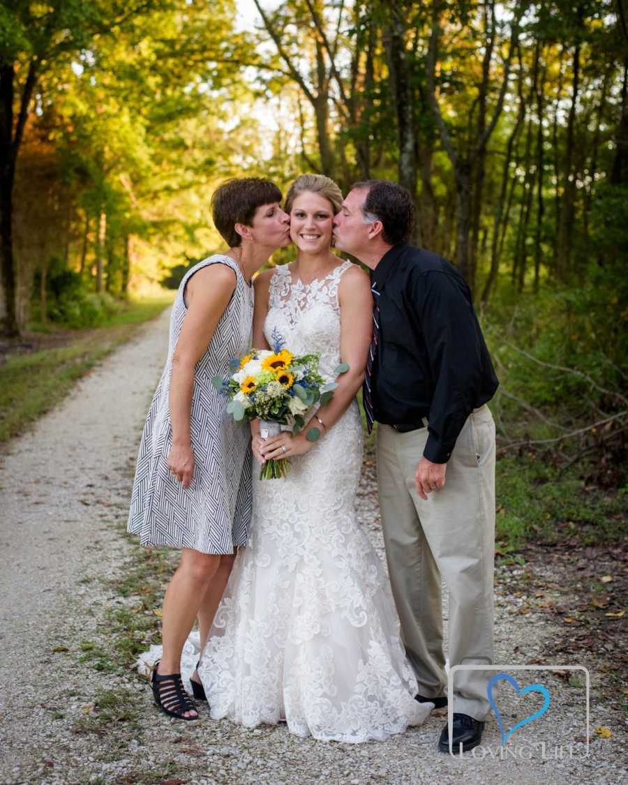 Woman in wedding gown stands with late fiancee's parents kissing her cheek