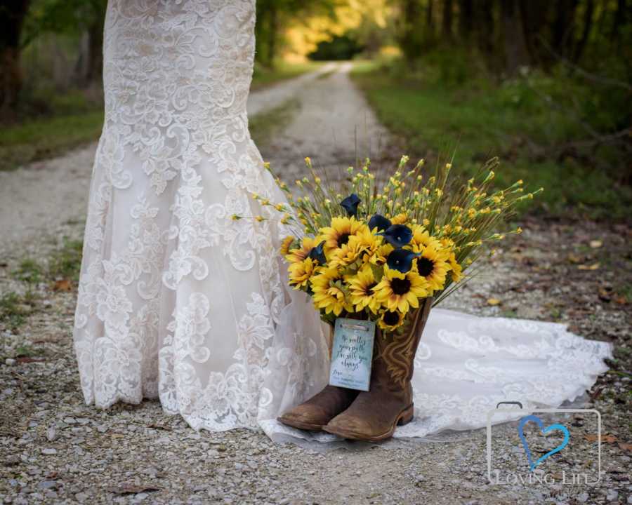 Late man's cowboy boots with flowers in them sit on dirt path beside woman in wedding gown