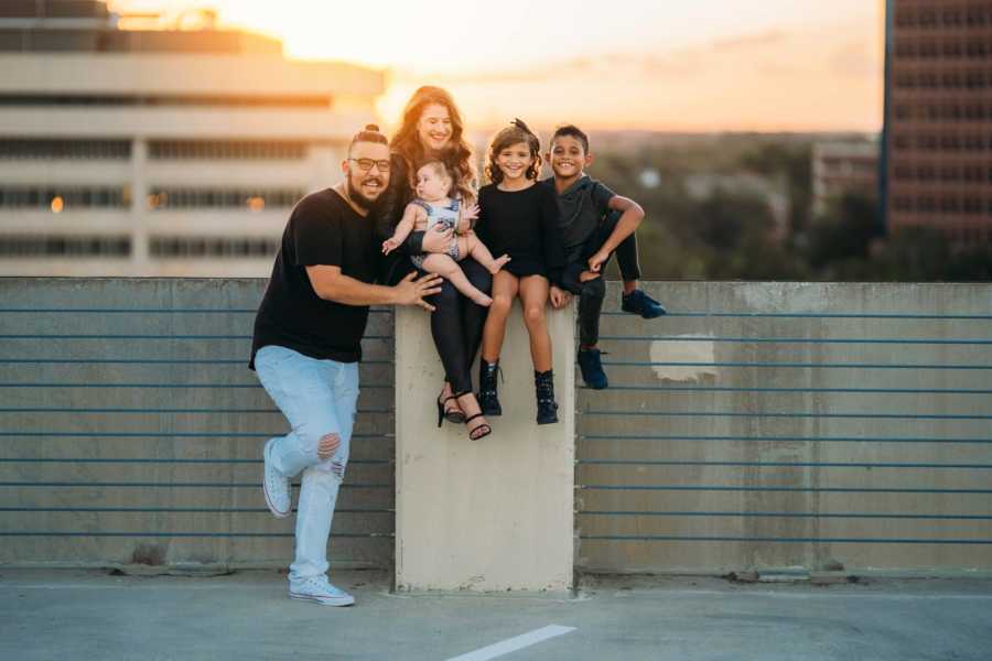 Woman sits on pillar of parking garage with her children and her boyfriend's daughter beside boyfriend