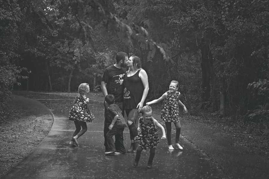Husband and wife kiss on street with their four kids dancing in rain