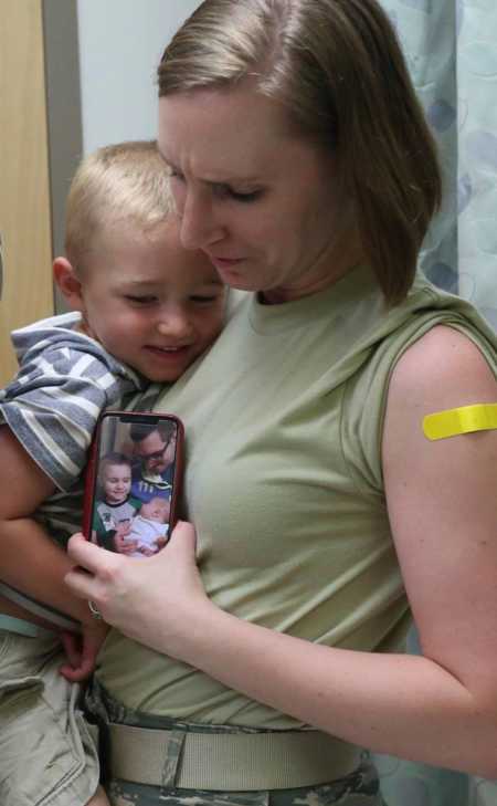Mother with band aid on arm from flu shot holding her son and phone with picture of husband and sons