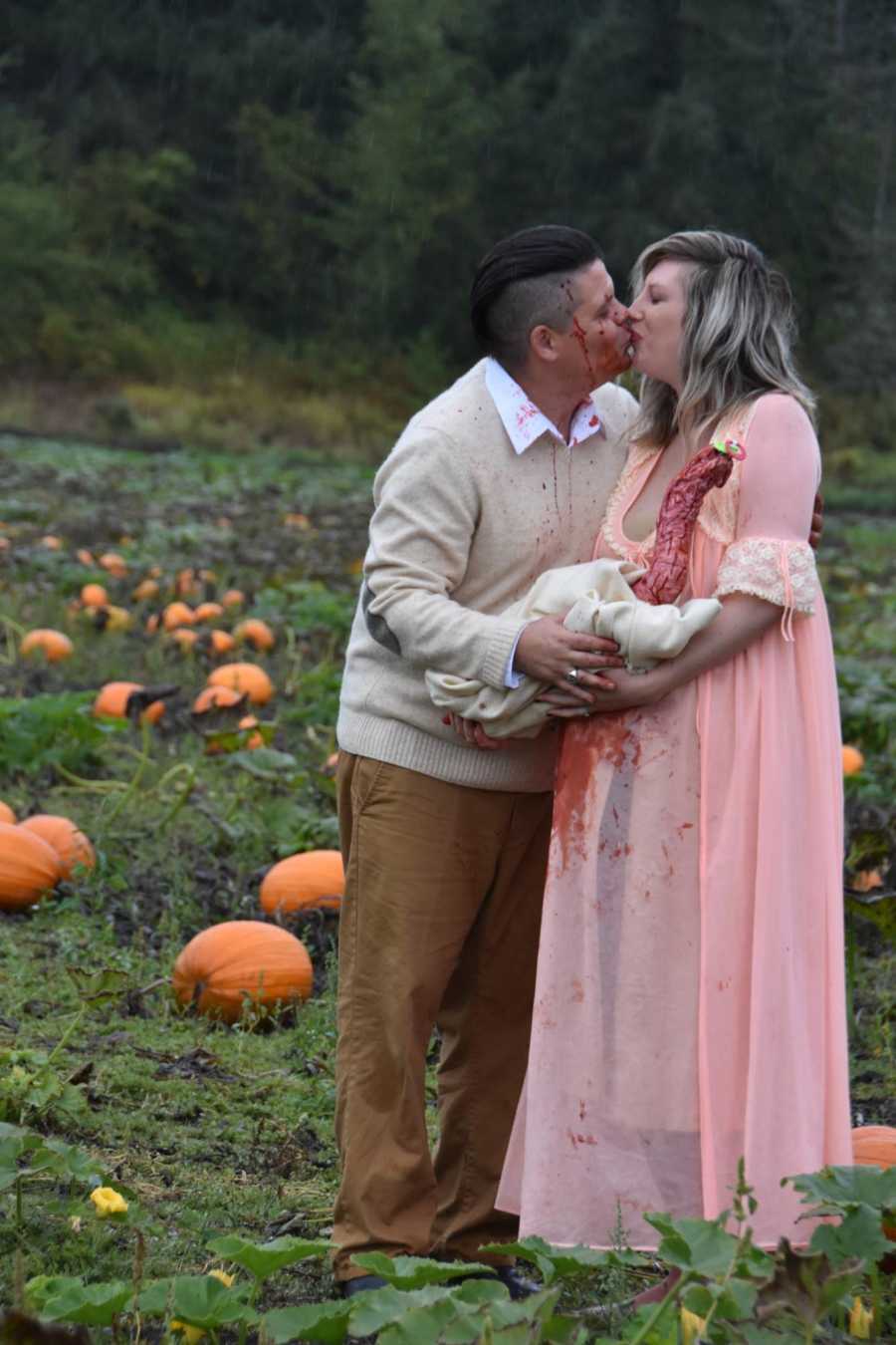 Husband and wife kiss as they hold bloody creature that came out of wife's stomach in pumpkin patch