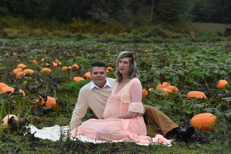 Pregnant woman sits on blanket with husband in pumpkin patch for pregnancy shoot