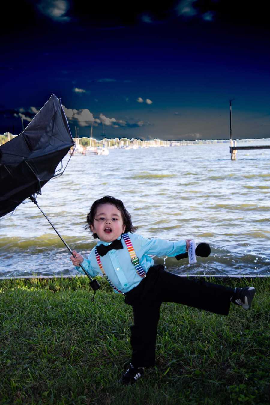 Toddler standing outside dressed as weather man holding microphone and umbrella near body of water