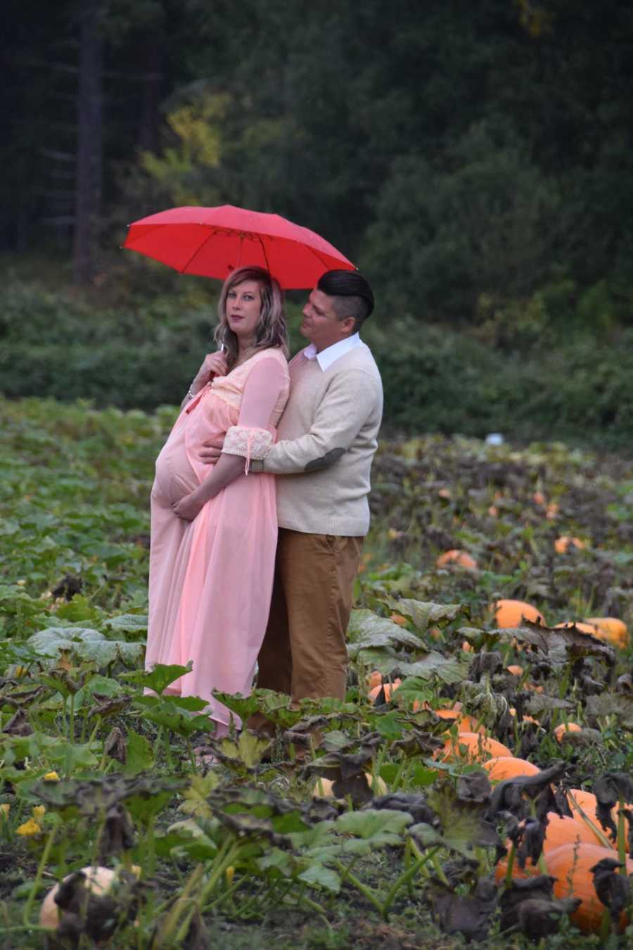 Pregnant woman stands holding umbrella in pumpkin patch with husband holding her from behind
