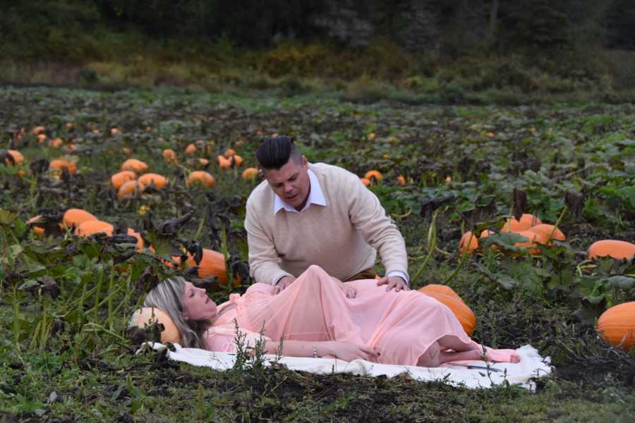 Pregnant woman lays on blanket in pumpkin patch with something protruding her stomach with husband watching