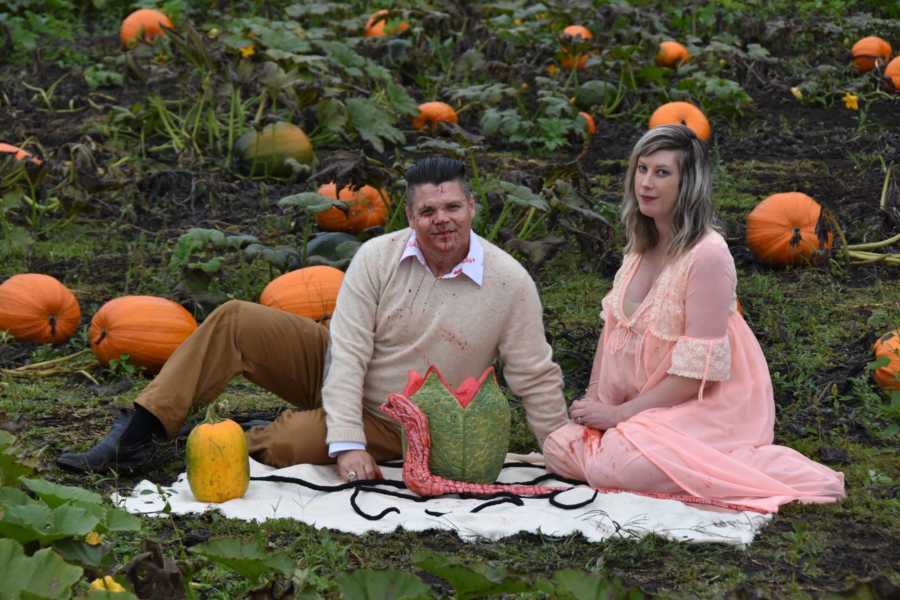 Husband and wife sit on blanket in pumpkin patch with bloody creature that came out of wife's stomach