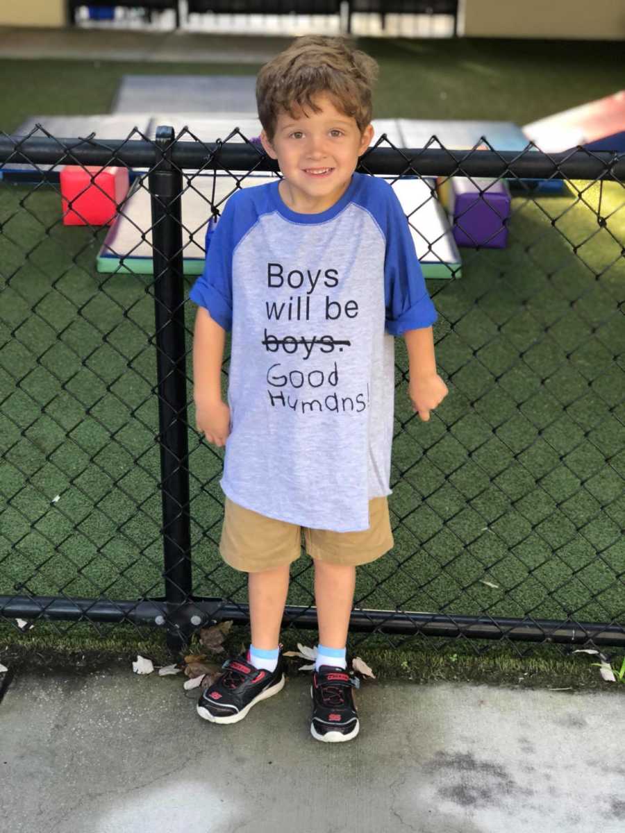 Little boy who was teased for playing with doll smiles in front of fence wearing shirt that says, "Boys will be good humans"