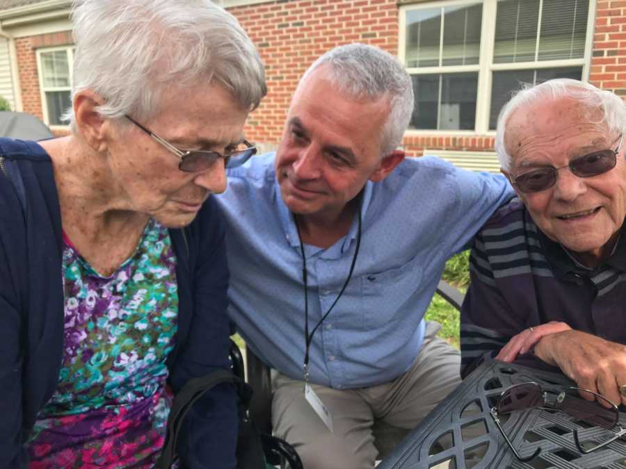 Man who was a foreign exchange student as a child sits with arm around his host with dementia and her husband