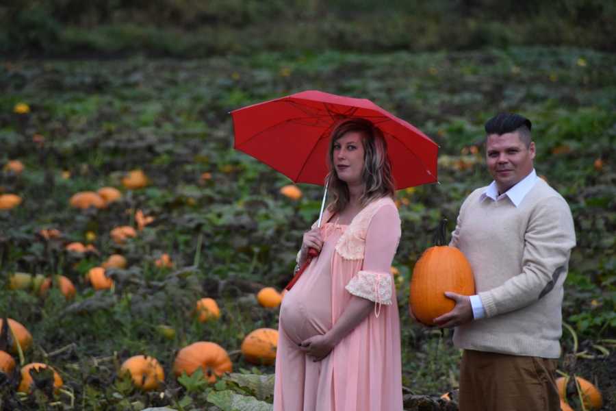 Pregnant woman holds red umbrella in pumpkin patch while husband is behind her holding pumpkin