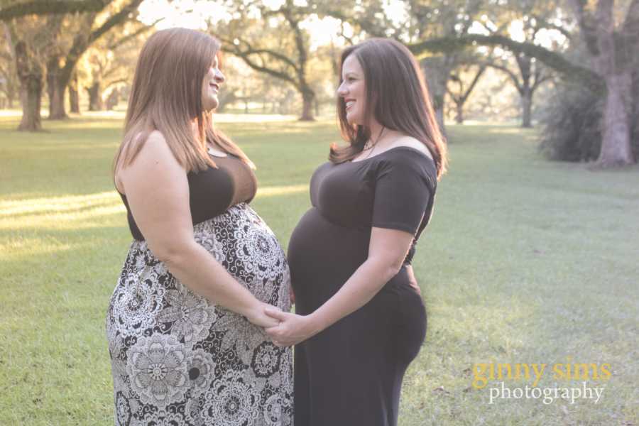 Pregnant wives hold hands and smile at each other while their stomachs touch