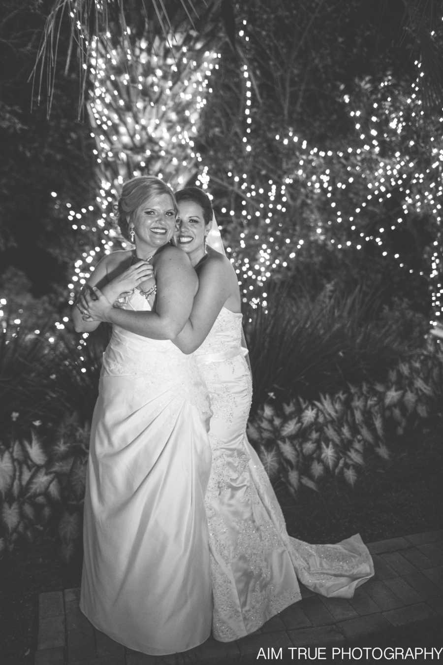 Wives hugging each other on their wedding day in wedding gowns beside tree wrapped in lights