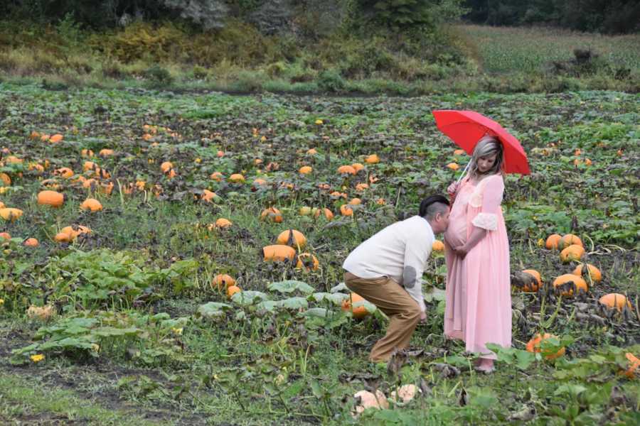Pregnant woman holds red umbrella in pumpkin patch while husband kisses her stomach
