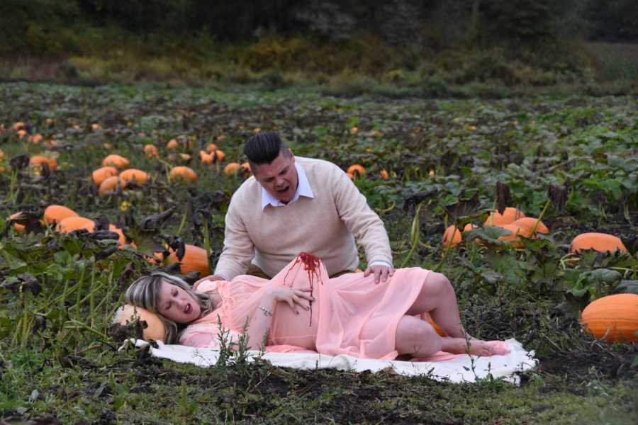 Pregnant woman lays on blanket in pumpkin patch while blood covers her dress and husband sits beside her