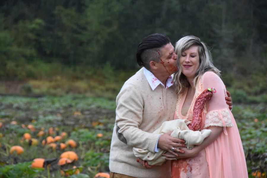 Husband kisses wife's cheek as they hold bloody creature that came out of her stomach in pumpkin patch