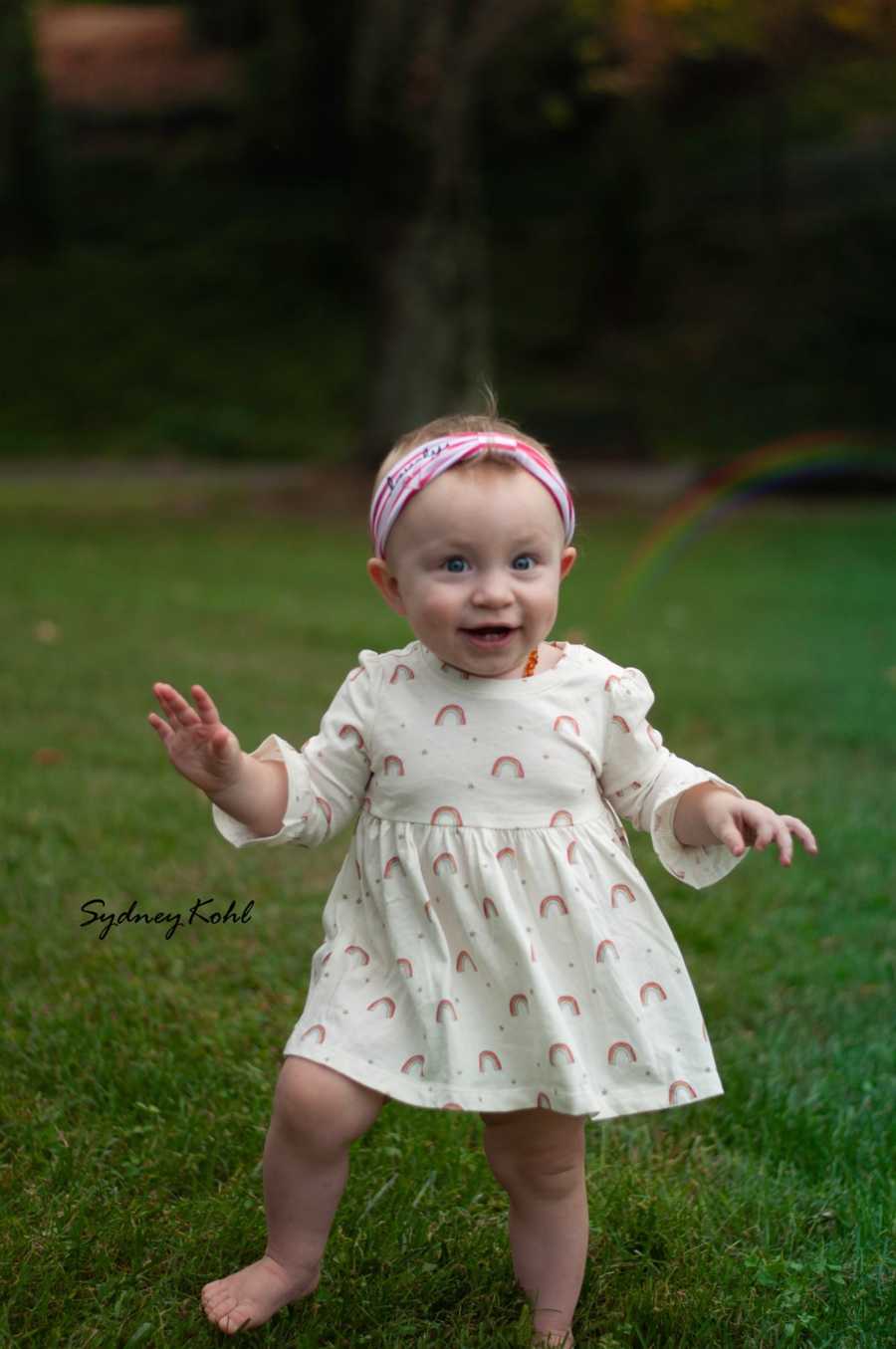 Little girl in rainbow dress stands smiling in grass whose mother lost younger sibling