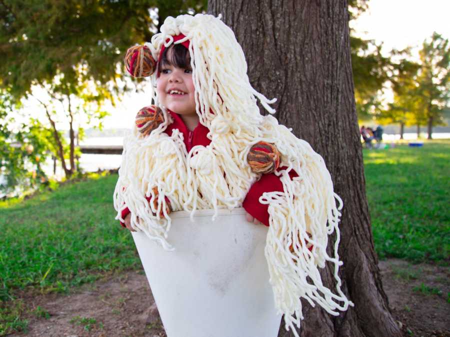 Toddler stands outside beside tree dressed as bowl of spaghetti and meatballs