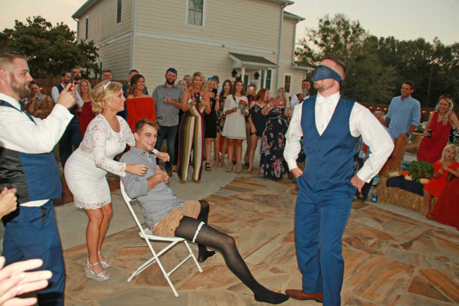 Groom blindfolded as bride stands behind chair where man sits with garter on