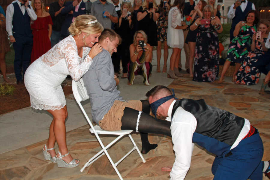 Blindfolded groom takes garter off of man that sits in front of bride who is laughing