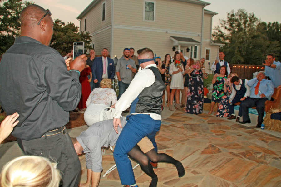 Blindfolded groom stands straddled around man with garter on who he thinks is his bride