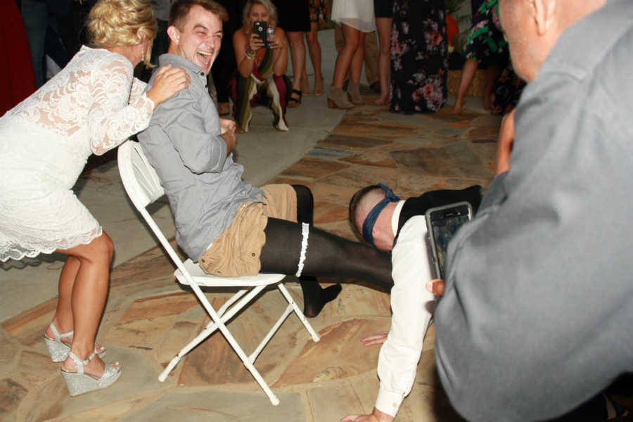 Bride stands behind man who is sitting in chair laughing as blindfolded groom takes garter off of him