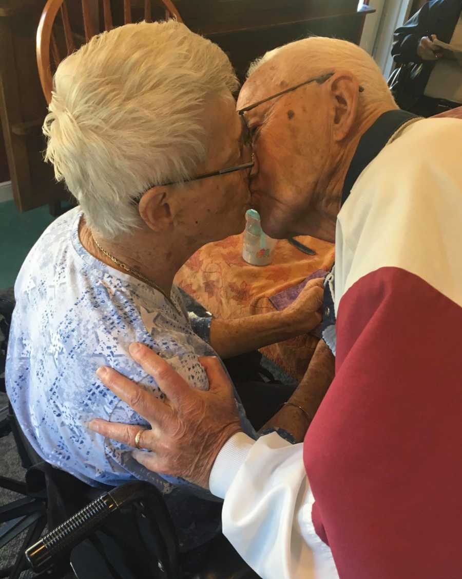 Woman with dementia sits in wheelchair while husband leans over to kiss her