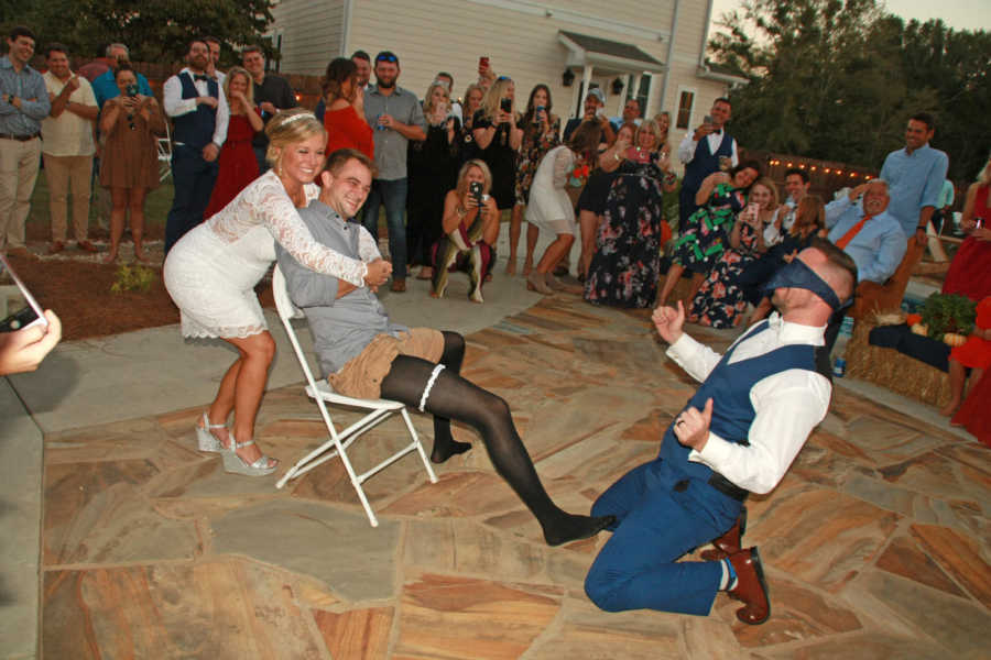 Bride stands behind man sitting in chair with garter on who is sitting in front of blindfolded groom