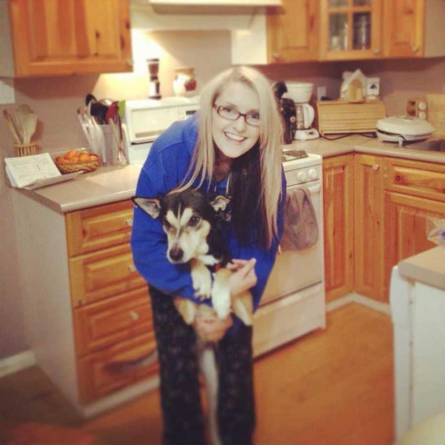 Woman stands in kitchen smiling while holding her dog that stands on its back legs