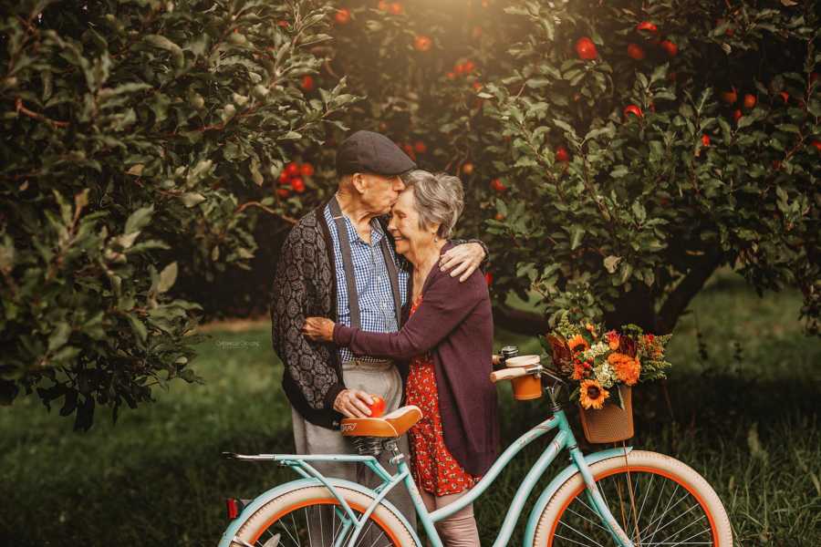 Elderly couple stand beside bike hugging while husband kisses wife's forehead