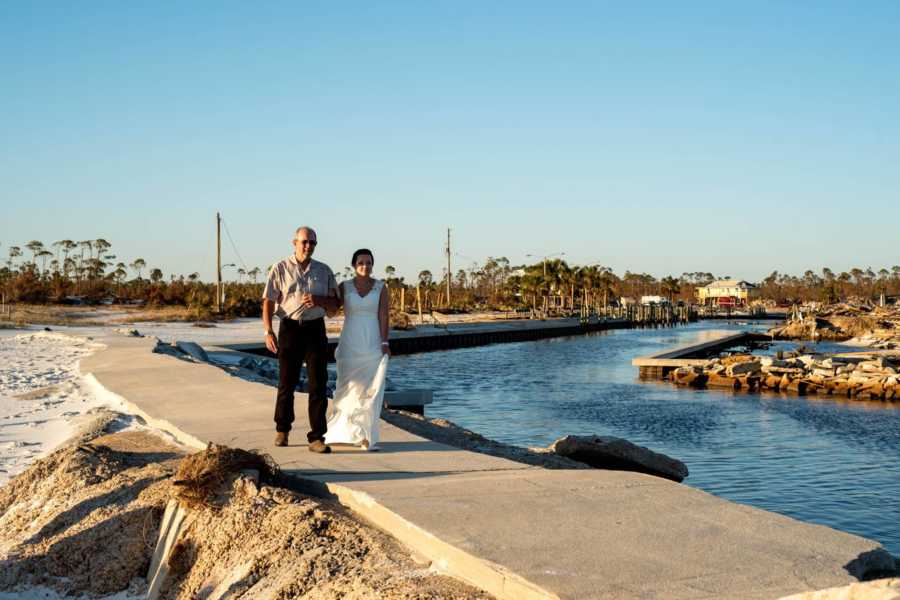 Bride walks arm in arm with father on sidewalk beside body of water