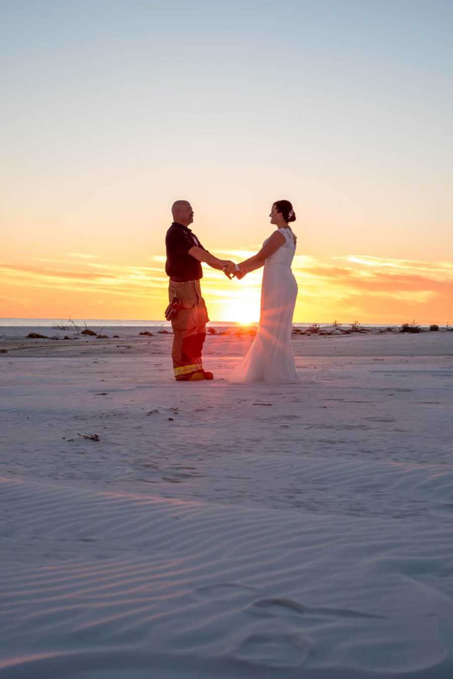 Bride and fireman groom holding hands on beach at sunset 