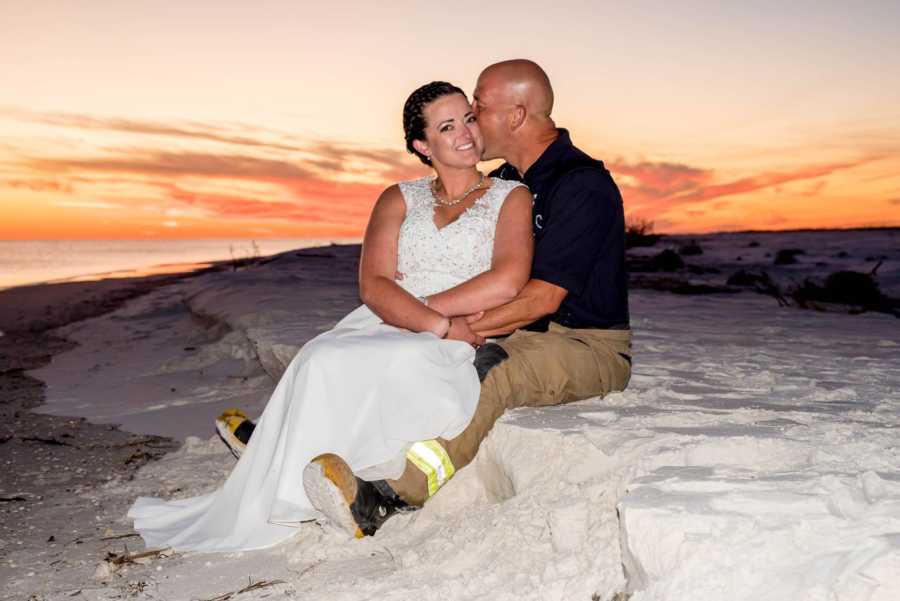 Bride and groom sit in sand while groom kisses her cheek