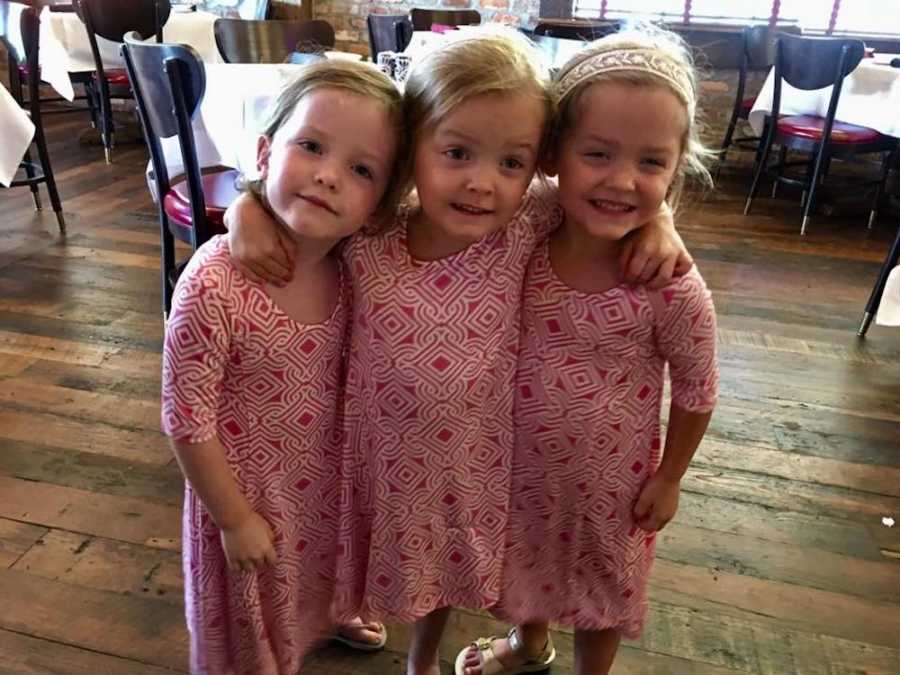 Triplet girls standing in matching dresses with arms around each other smiling