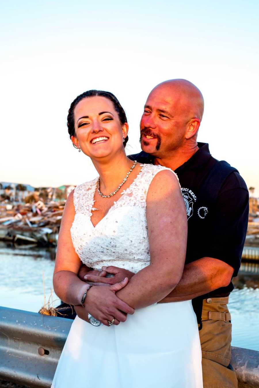 Groom holds bride from behind smiling near body of water