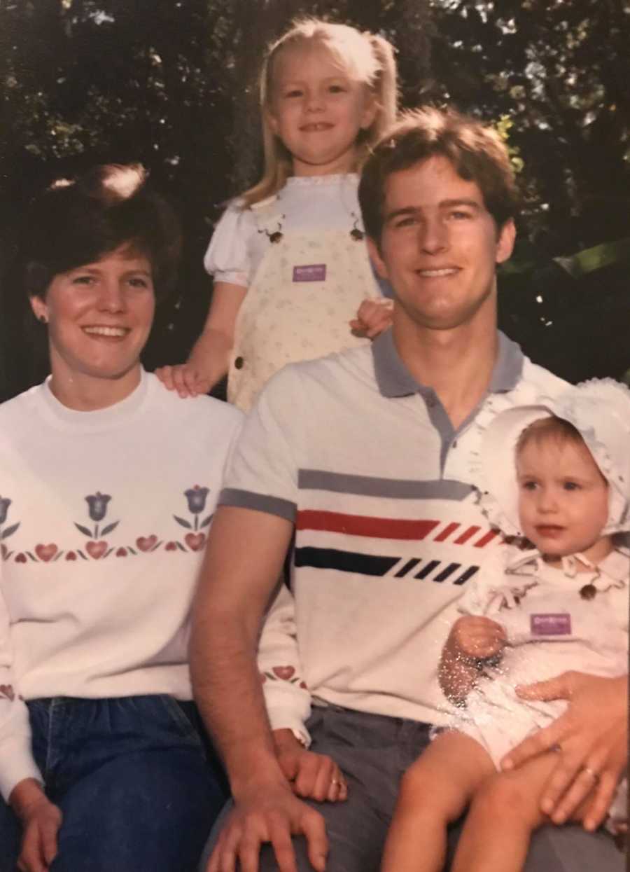 Husband and wife sit smiling with their first born standing behind them and youngest on father's lap