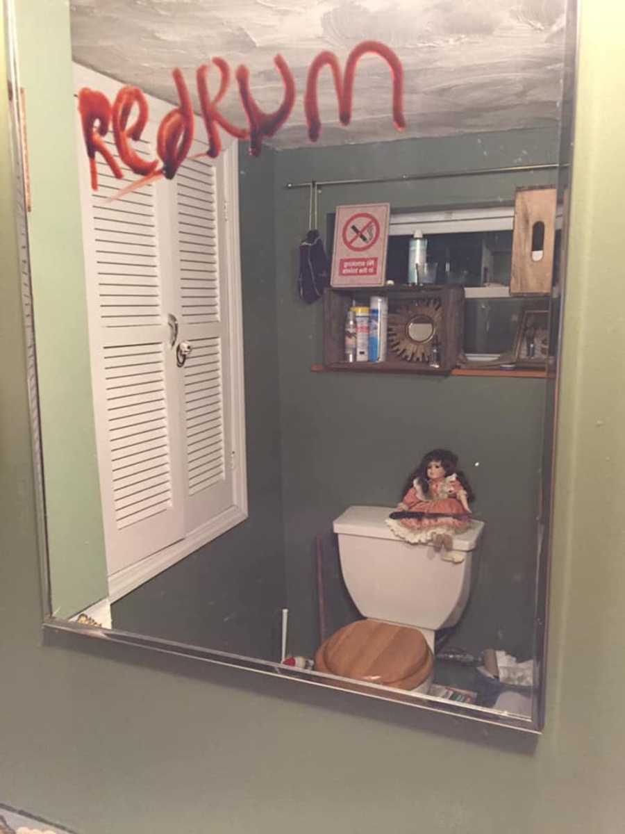 Redrum written in red on mirror with reflection of doll sitting on top of toilet