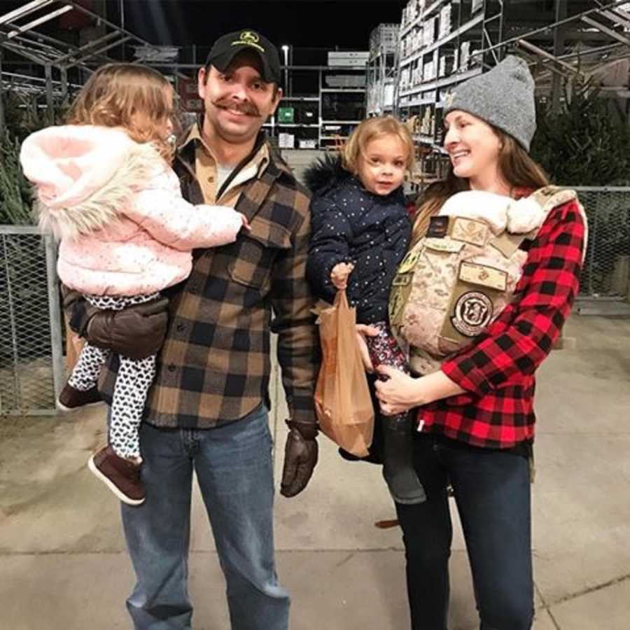 Husband with PTSD stands holding daughter beside wife who is holding their other two children