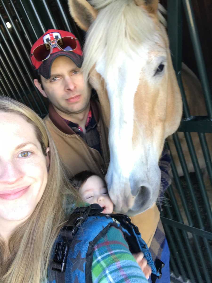 Wife smiles in selfie with daughter swaddled to her back and her husband with PTSD standing beside horse