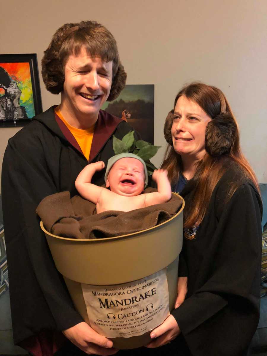 Husband and wife wearing earmuffs and Hogwarts robes stand holding crying baby in bucket that says, "Mandrake"