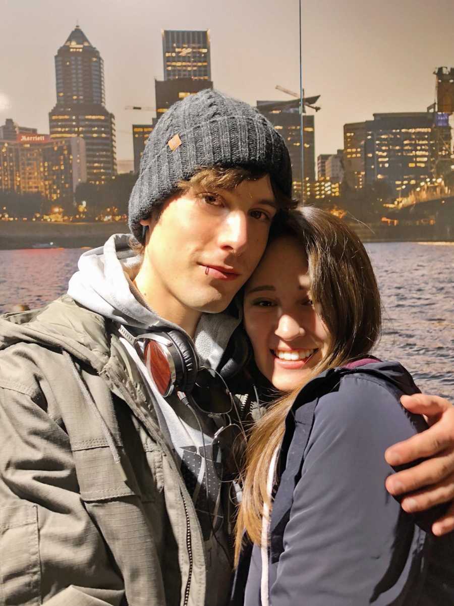 Young man who struggled with addiction stands with arm around girlfriend with body of water and skyline in background