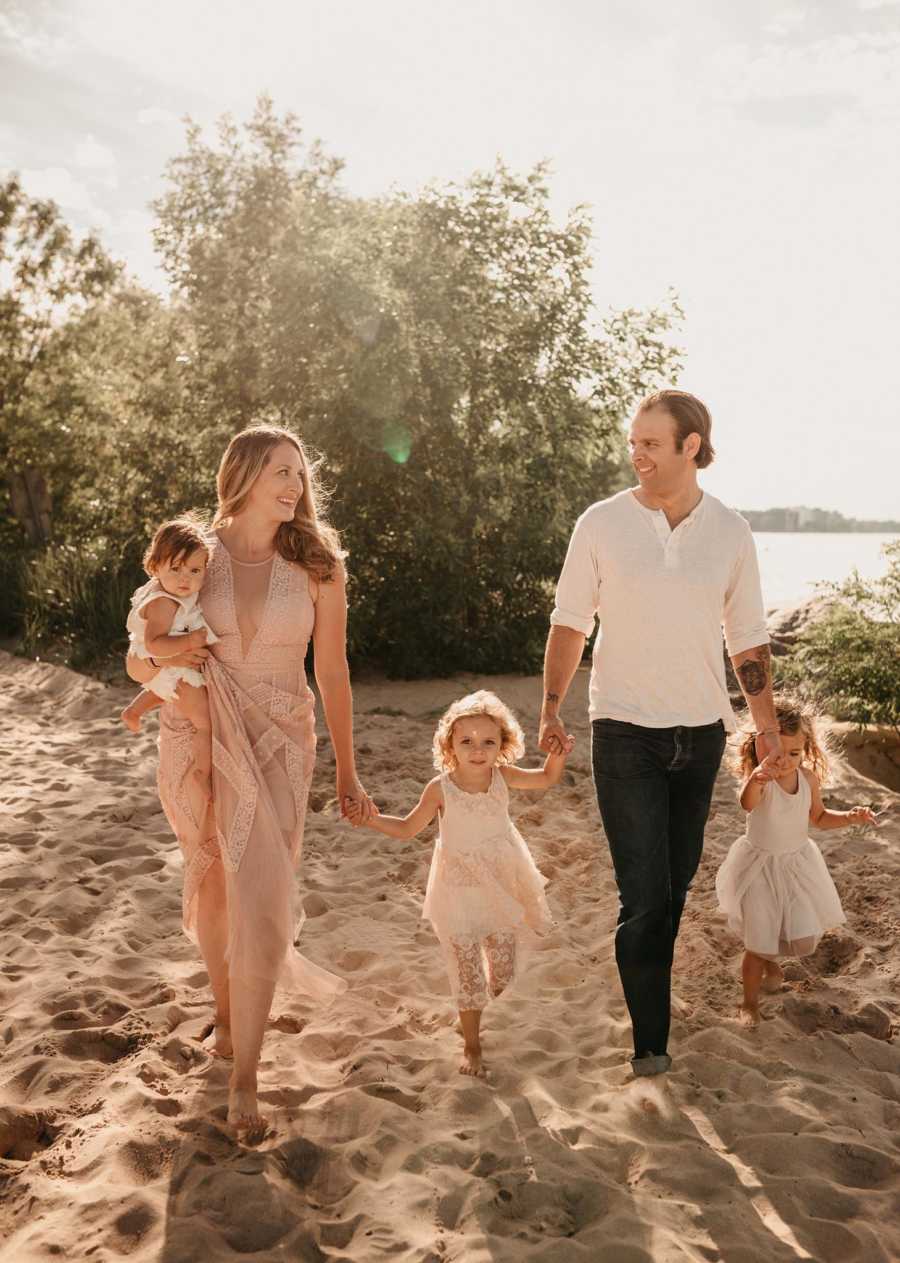 Husband and wife smile at each other as they walk along beach holding hands with their children 