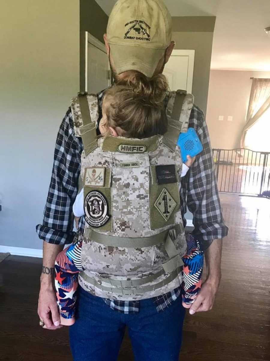 Father with PTSD from war stands in home with daughter swaddled to his chest in camo baby carrier